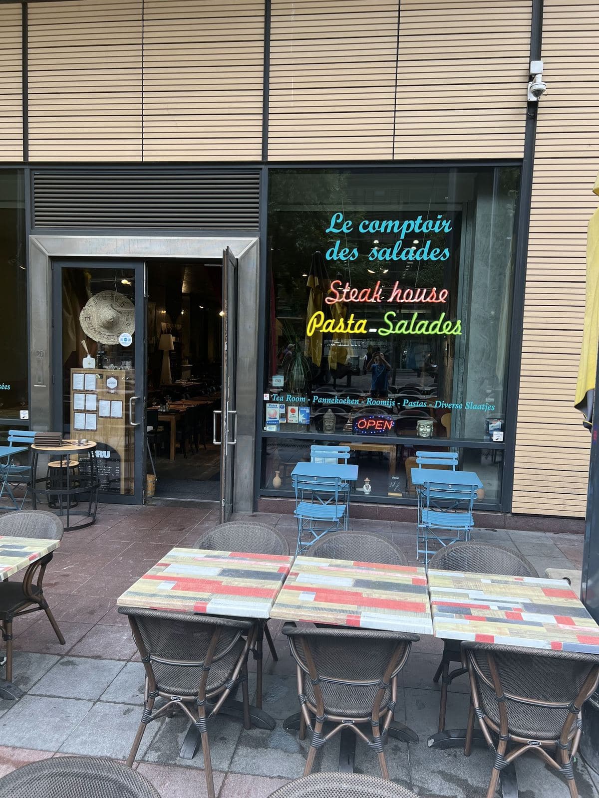 La façade place Victor Horta : les néons « Le comptoir des salades », « Steak house » et « Pasta – Salades » en vitrine, la terrasse et ses tables colorées devant