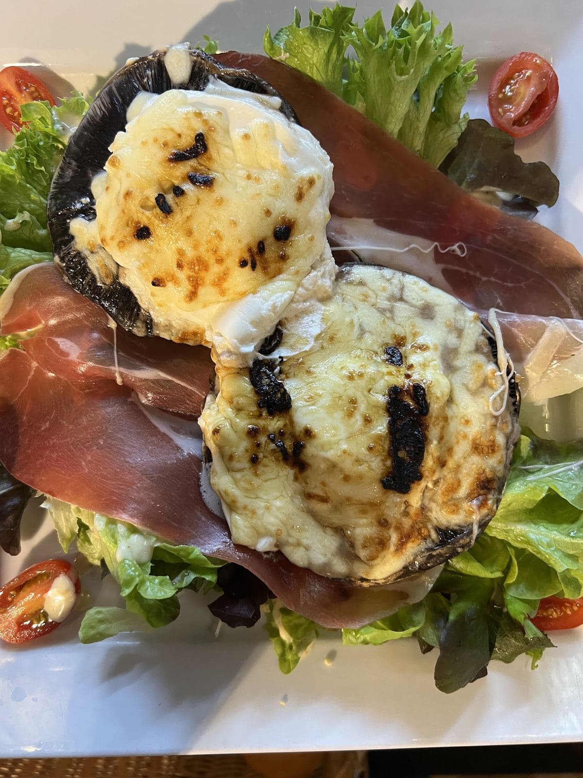 Deux toasts de chèvre gratiné posés sur du jambon cru et une salade verte, avec des tomates cerise