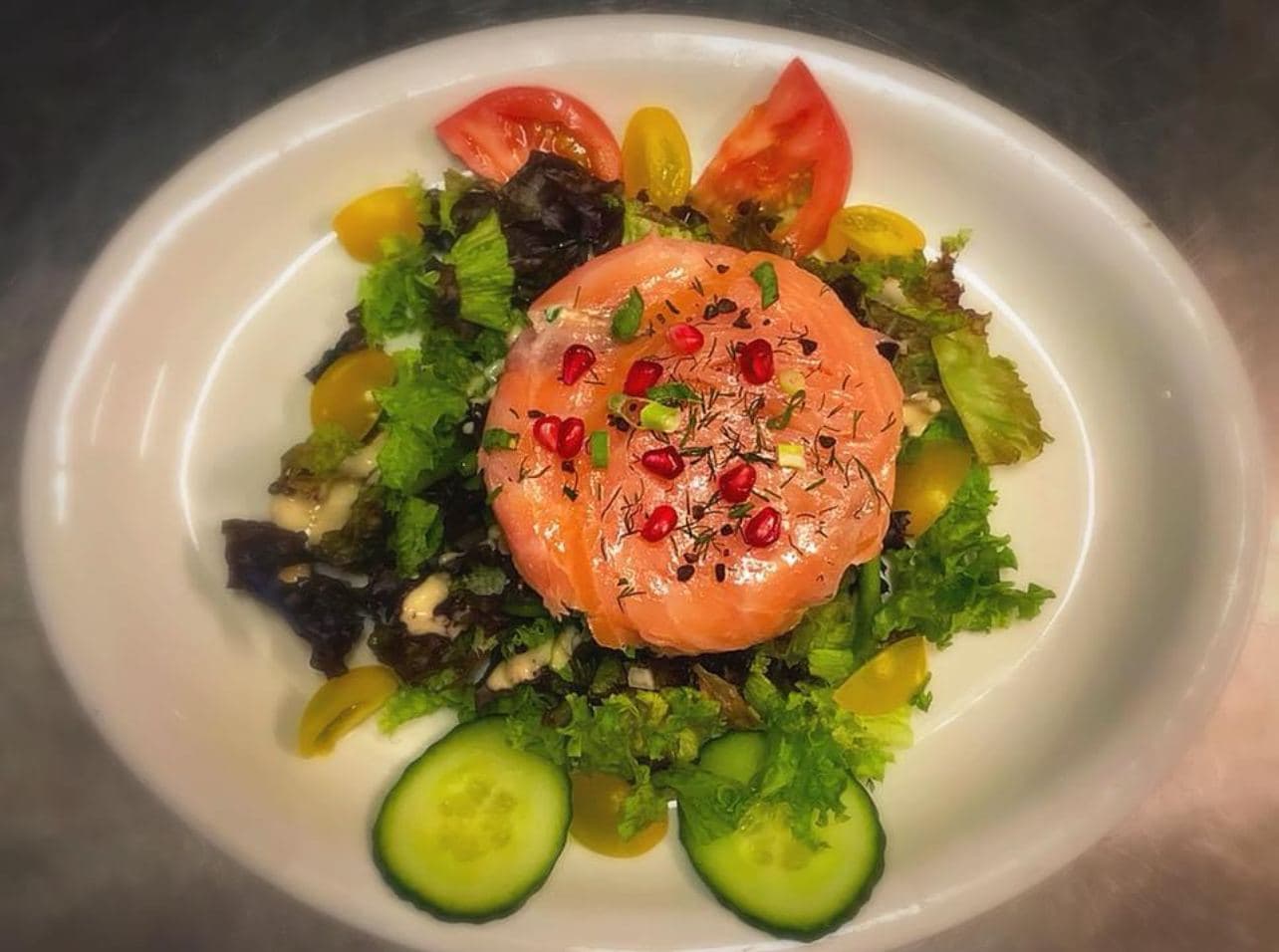 Un tartare de saumon parsemé de grenade, au centre d'une salade verte bordée de tomates et de rondelles de concombre