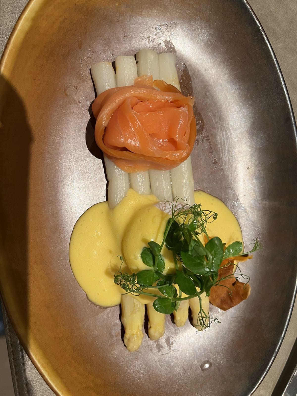 Des asperges blanches, une rose de saumon fumé et une sauce jaune, sur une assiette ovale cuivrée