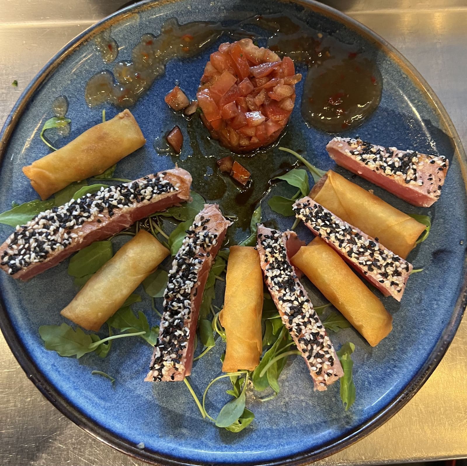Des tranches de thon rouge en croûte de sésame noir et blanc, en étoile avec des nems, autour d'un tartare de tomate, sur une assiette bleue